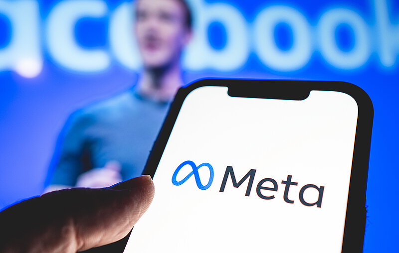 Ein Smartphone, auf dem das Metasymbol zu sehen ist, dahinter verschwommen Mark Zuckerberg