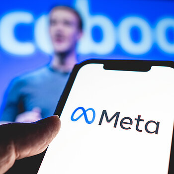 Ein Smartphone, auf dem das Metasymbol zu sehen ist, dahinter verschwommen Mark Zuckerberg