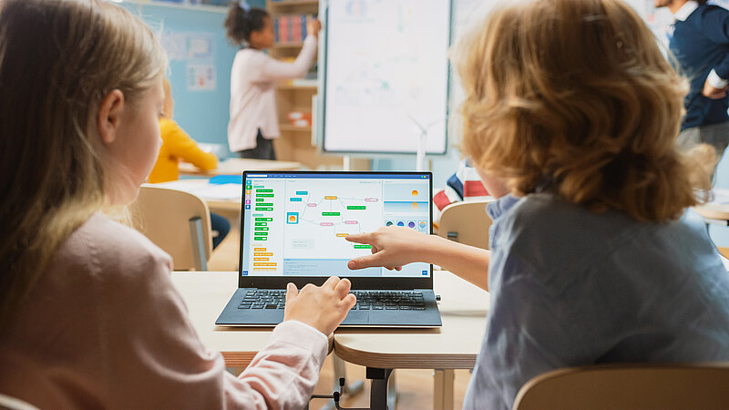 Zwei Kinder von hinten, die an ihren Schulschreibtischen sitzen und auf ein Diagramm auf einem Laptop zeigen