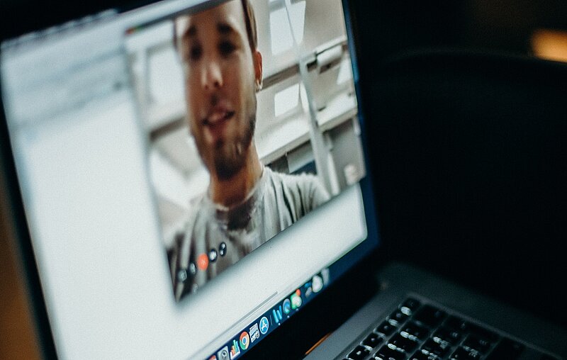 Ein Laptop im Dunkeln, auf dessen Bildschirm das Gesicht eines Mannes während einer Videokonferenz zu sehen ist