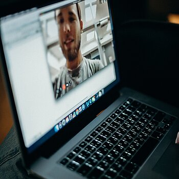 Ein Laptop im Dunkeln, auf dessen Bildschirm das Gesicht eines Mannes während einer Videokonferenz zu sehen ist