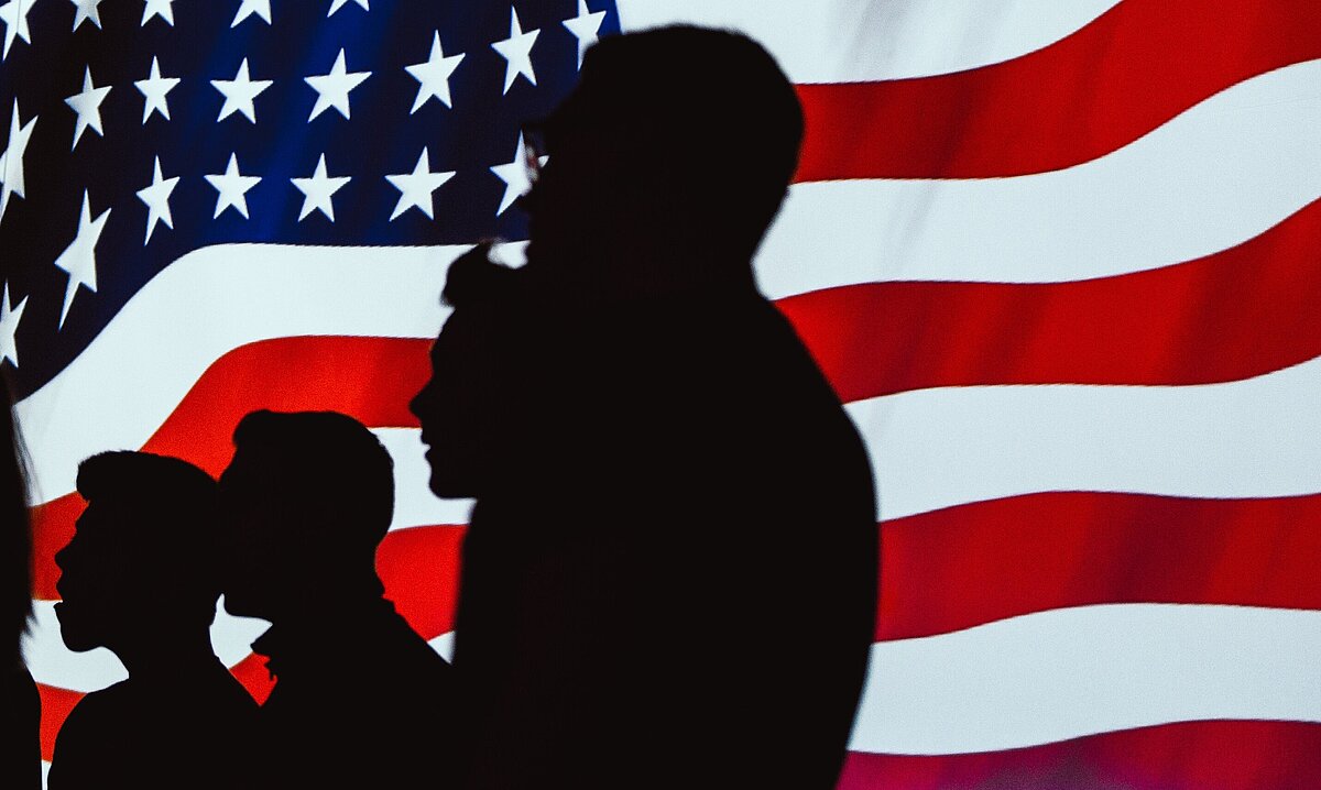 Die US-Flagge im Dunkeln, von hinten Beleuchtet, mit vier Menschen als schwarze Schatten im Vordergrund